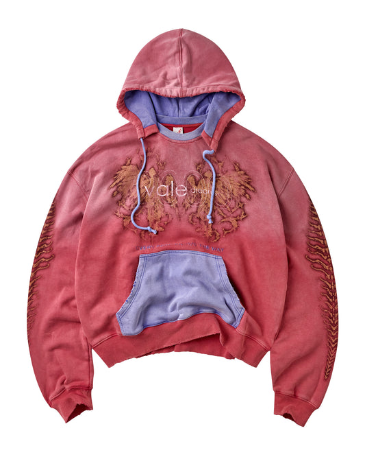 A cropped pullover hoodie in a washed, faded red-pink color, featuring a light purple/lavender hood lining, drawstrings, and a matching front kangaroo pocket. The chest has a graphic of two golden-brown angels, and the sleeves have a design pattern.